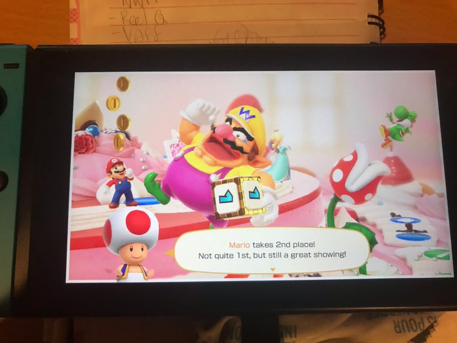 photo of nintendo switch on a notebook and lens cleaning fabric. the game is mario party, with wario, mario, yoshi and rosalina posing on peach's birthday cake. wario in the front holds a custom dice block and also has no teeth. the text from toad reads 'mario takes 2nd place! not quite 1st, but still a great showing!'